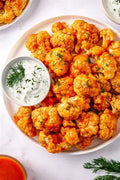A top-down view of buffalo cauliflower on a white plate, served with a small bowl of white ranch dressing and garnished with fresh dill, on a white marble surface.